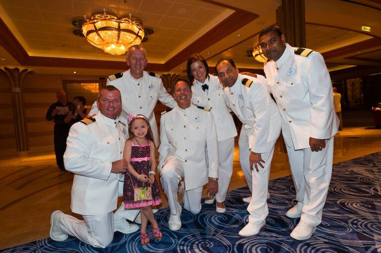 Young girl in a vibrant dress smiling with a group of friendly cruise ship officers in white uniforms onboard a luxury cruise liner, symbolizing family-friendly cruise experience