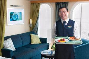 Smiling waiter serving breakfast in a luxury cruise ship suite with elegant interior design and ocean view through the porthole.