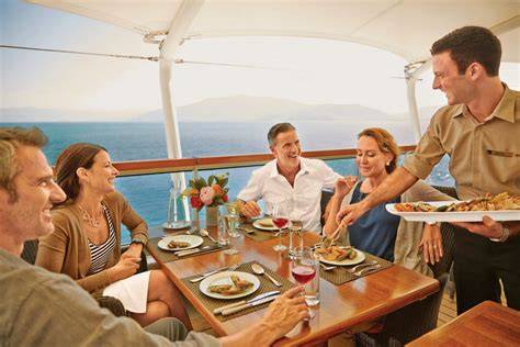 Passengers enjoying drinks at an outdoor bar on a cruise ship, with a bartender shaking a cocktail and the ocean in the background.