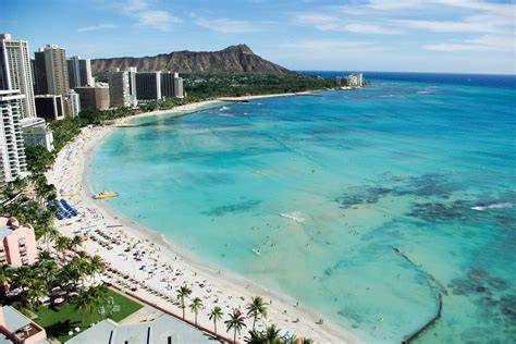 A panoramic view of a stunning tropical beach with clear turquoise waters, lined with high-rise buildings, and a distant mountain backdrop.