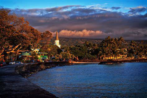 Coastal village with a church, lush greenery, and calm water under a dramatic sunset sky.