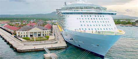 Large cruise ship docked at a scenic port with historic buildings and a coastal landscape in the background.