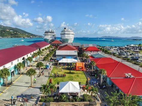 A vibrant cruise port in the Caribbean featuring two large cruise ships docked. The scene includes colorful buildings with red roofs, green palm trees, and people enjoying the sunny day, representing a bustling maritime travel destination.