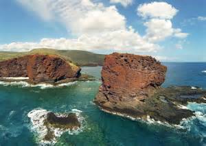 Rocky coastal landscape with large red rock formations and clear blue ocean water under a partly cloudy sky.