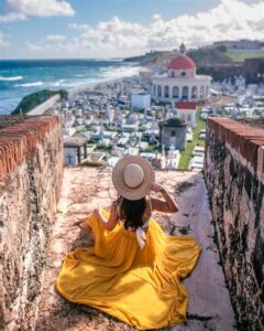 Woman in a yellow dress sitting on a historic wall overlooking a coastal town and ocean