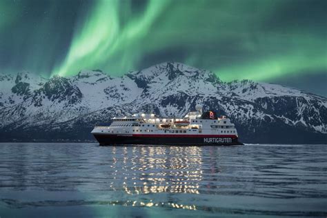 Cruise ship sailing under the Northern Lights with snow-covered mountains in the background.