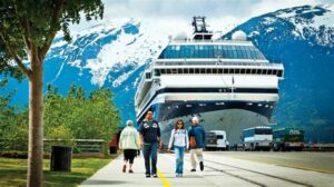 Passengers walking towards a cruise ship docked in an Alaskan port with snow-covered mountains in the background.